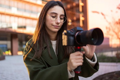 A woman in a green coat is focused on using a camera during a sunset outdoor shoot The background