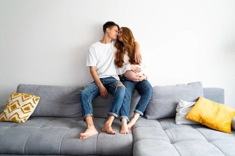 An expectant couple seated on a comfortable gray sofa, lovingly embracing each other. They are