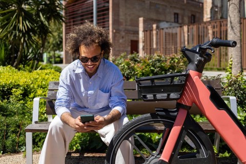 Afro man sitting on a park bench, smiling while looking at his smartphone. A bicycle is parked