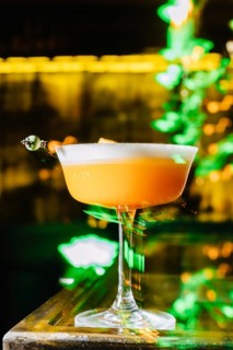 A vibrant peach cocktail with a frothy top, elegantly served in a coupe glass. The cocktail sits on