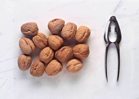 A close-up image of walnuts paired with a metal nutcracker on a white marble surface. The