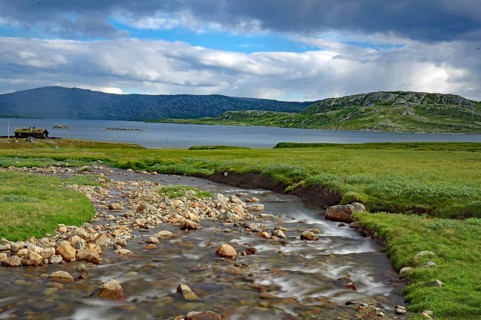 A small river flows through green fields under a cloudy sky with mountains in the background, Fell,