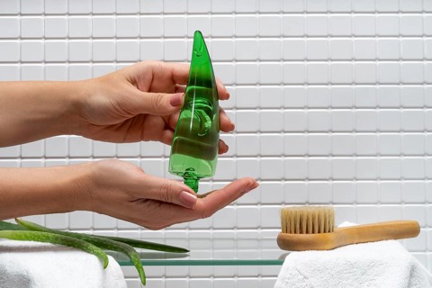 Cropped unrecognizable woman dispenses aloe vera gel onto her hand against a tiled bathroom