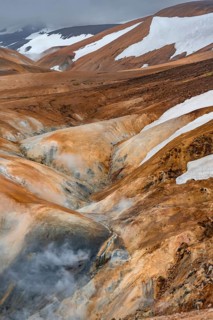 Steaming stream between colourful rhyolite mountains with snowfields, Hveradalir geothermal area,