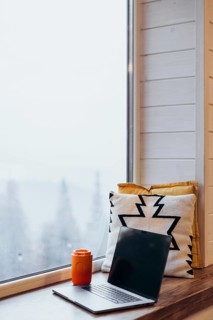 A laptop open next to a bright orange thermos cup on a windowsill, with snowy scenery outside. A