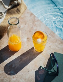 Refreshing glass of fresh orange juice placed on a sunlit table beside a pool. The ambient scene