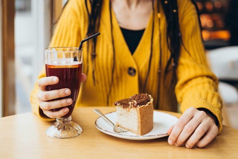 A close-up of a person in a yellow sweater holding a glass with a drink, and a plate with a slice