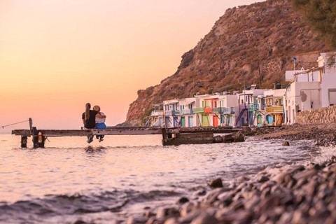 A couple seated on a wooden dock overlooks the vibrant, colorful houses of Klima, Milos Island,