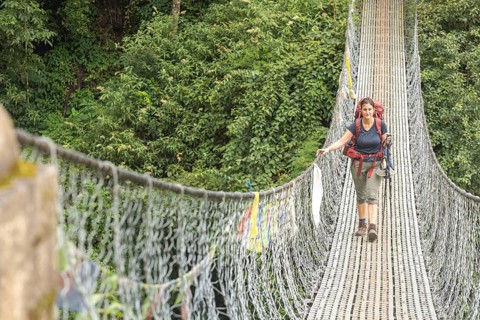 A woman trekker with a backpack crosses a suspension bridge surrounded by lush greenery in the