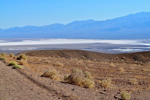 A barren landscape, with a salt pan in the foreground, surrounded by mountains under a blue sky,