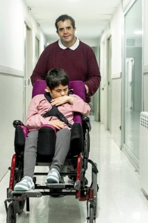 A man assists a woman in a wheelchair down a hallway in a mental health residence. The setting is