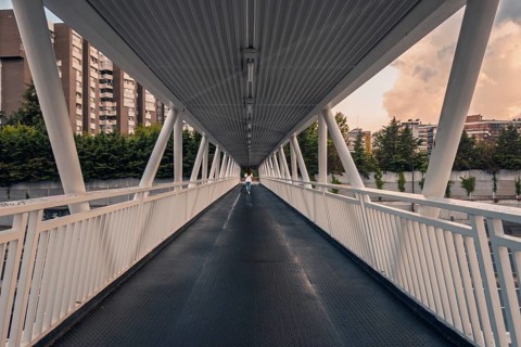 Back view of unrecognizable woman walking on a modern pedestrian bridge in Madrid. The sky is
