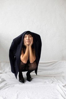 A thoughtful Asian woman poses creatively, with a jacket draped over her shoulders, against a