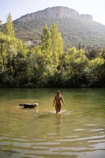 A woman and her dog enjoy a serene day at a lush green lake, surrounded by trees and a rocky hill