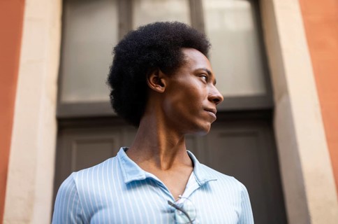 A non-binary person stands outside a modern building, gazing thoughtfully into the distance. The