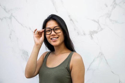 Smiling Chinese woman stands near a marble wall in a city, adjusting her glasses while enjoying the
