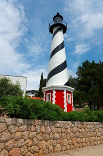 A black and white lighthouse in front of a stone wall, framed by green plants under a clear sky,