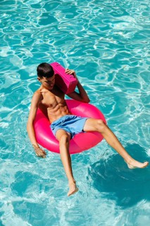 A teenage boy lounges on a bright pink pool float in clear blue water, holding a vibrant pink