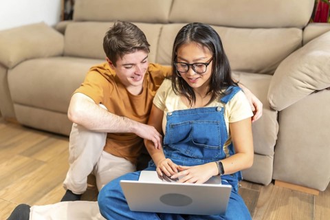 Happy multiethnic couple relaxing on floor at home, using laptop and browsing internet, enjoying
