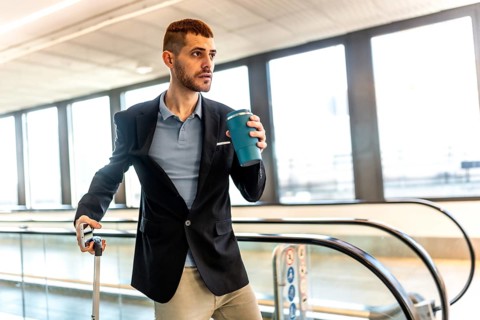 Young businessman drinking coffee while waiting at the airport terminal, holding his suitcase and