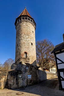 A stone tower under a blue sky, flanked by half-timbered houses and a cobbled path, watchtower of