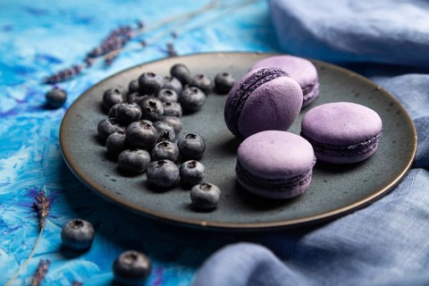 Purple macarons or macaroons cakes with blueberries on ceramic plate on a blue concrete background