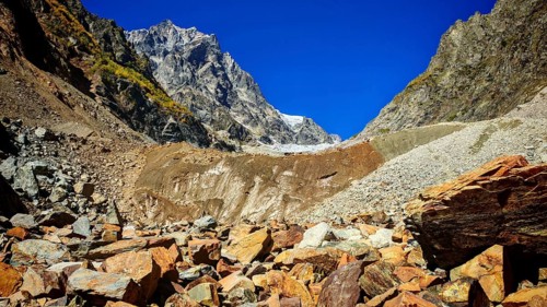 Red debris leads to steep mountain slopes under clear blue skies, hiking to Chalaadi Glacier,