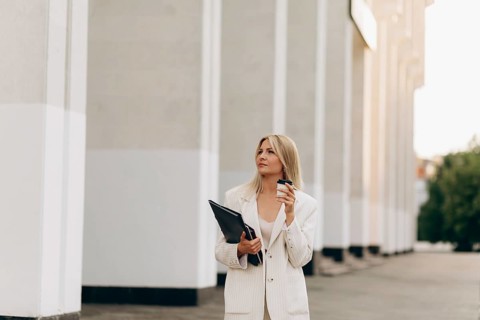 A businesswoman in a stylish white suit holds a coffee cup and folder, standing outdoors. The