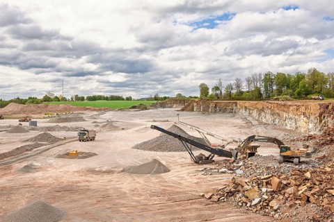 An open-pit quarry with piles of crushed stone and gravel, featuring heavy machinery including an