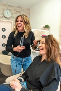 Happy hairdresser using a flat iron on a smiling female client in a busy modern salon, sharing