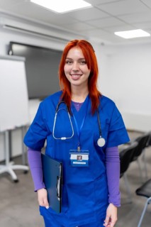 Young woman with red hair wearing blue scrubs and stethoscope, holding a clipboard and id badge,