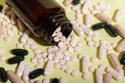 A variety of pills and capsules are scattered from a dark brown bottle onto a light green surface,