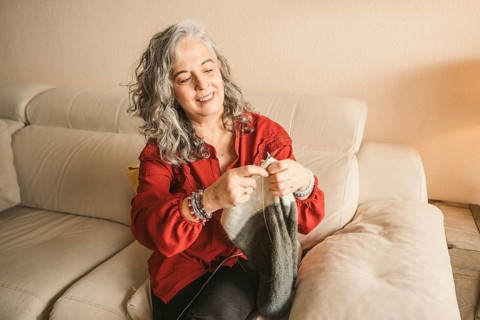 A joyful woman with gray hair knits a scarf while sitting on a comfortable beige couch. She wears a