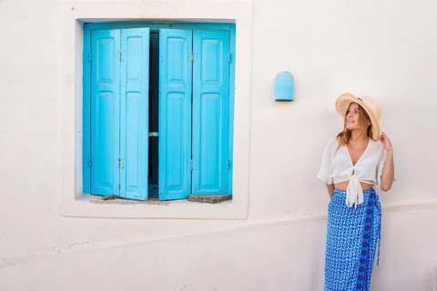 A woman in a straw hat and white blouse stands by a vibrant blue shutter in Trypiti, Milos Island,