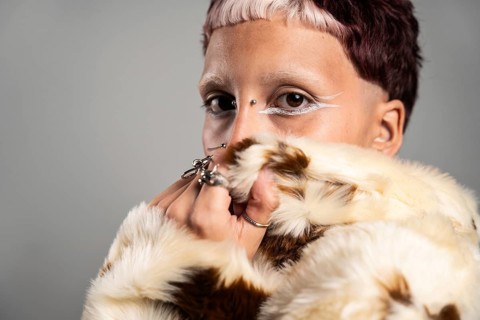 Close-up of a woman with striking makeup, bold rings, and a fur jacket. The expressive eyes and