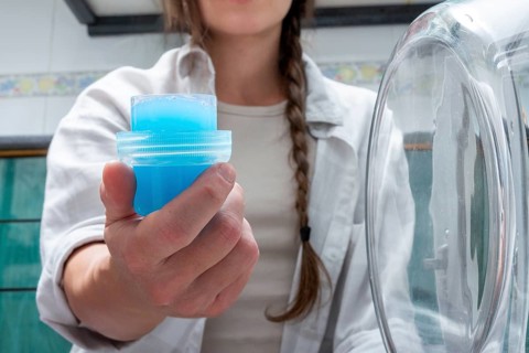 A Gen-Z girl holds a cap full of bright blue liquid detergent next to a washing machine,