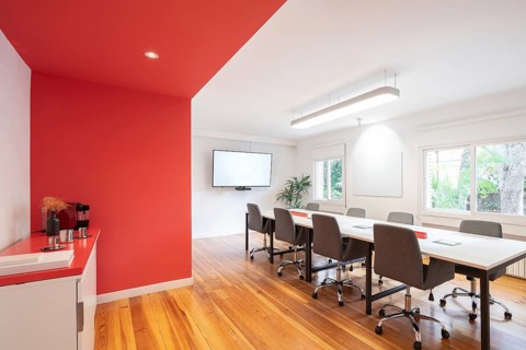 Bright meeting room in office with a long table, chairs, and a wall mounted screen. Red accents