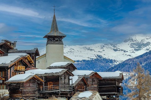 Old wooden houses with snow-covered roofs and the Saint Theodule church in the historic village
