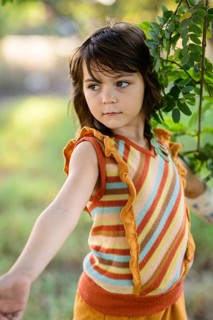 A young girl in a colorful striped top gazes thoughtfully beside a green tree branch. Her attire