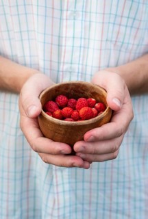 Man gently cradle a rustic wooden bowl filled with vibrant red strawberries. The blurred plaid