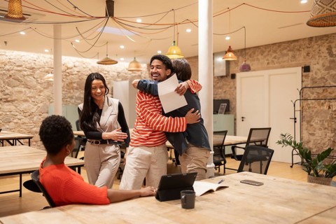 Four colleagues share a joyful moment in a stylish coworking space with exposed stone walls and