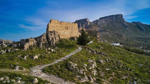 Drone shot, stone ruin on hill with path, embedded in mountainous landscape in front of blue sky,