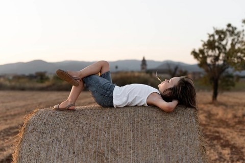 A boy relaxes on a hay bale in the countryside, surrounded by serene fields at sunset. The peaceful
