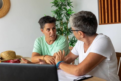 Two women discussing travel plans at home, with a laptop and summer hats on the table, depicting a