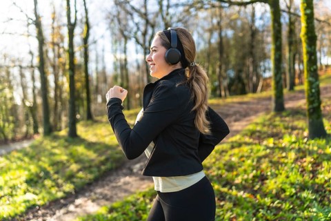 Woman jogging on a leafy park trail wearing headphones and sportswear, smiling as she enjoys a