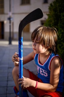 A young boy in a sporty outfit crouches while holding a blue hockey stick. The setting is outdoors