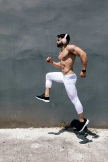 Fit man jogging energetically against a concrete wall wearing white leggings and black sneakers. He