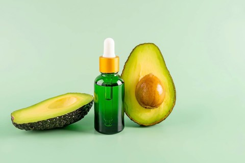 A green bottle of avocado oil with a dropper, placed between two fresh avocado halves on a light