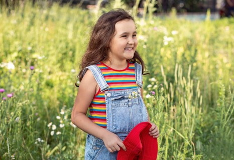 A cheerful little girl stands smiling in a sunlit meadow, holding a chamomile flower. Her playful