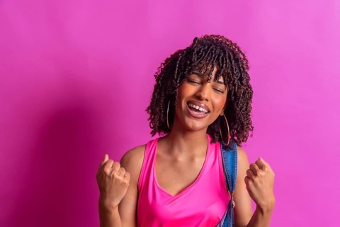 Young woman with curly hair, wearing a pink top and denim overalls, joyfully celebrating against a